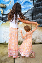 Lifestyle shot of a woman and child wearing matching Aria Spring Dress Sunrise Botanica Rayon, a bohemian-style clothing in soft peach-orange, dusty rose pink, and light lavender floral patterns, paired with a solid white crewneck top.