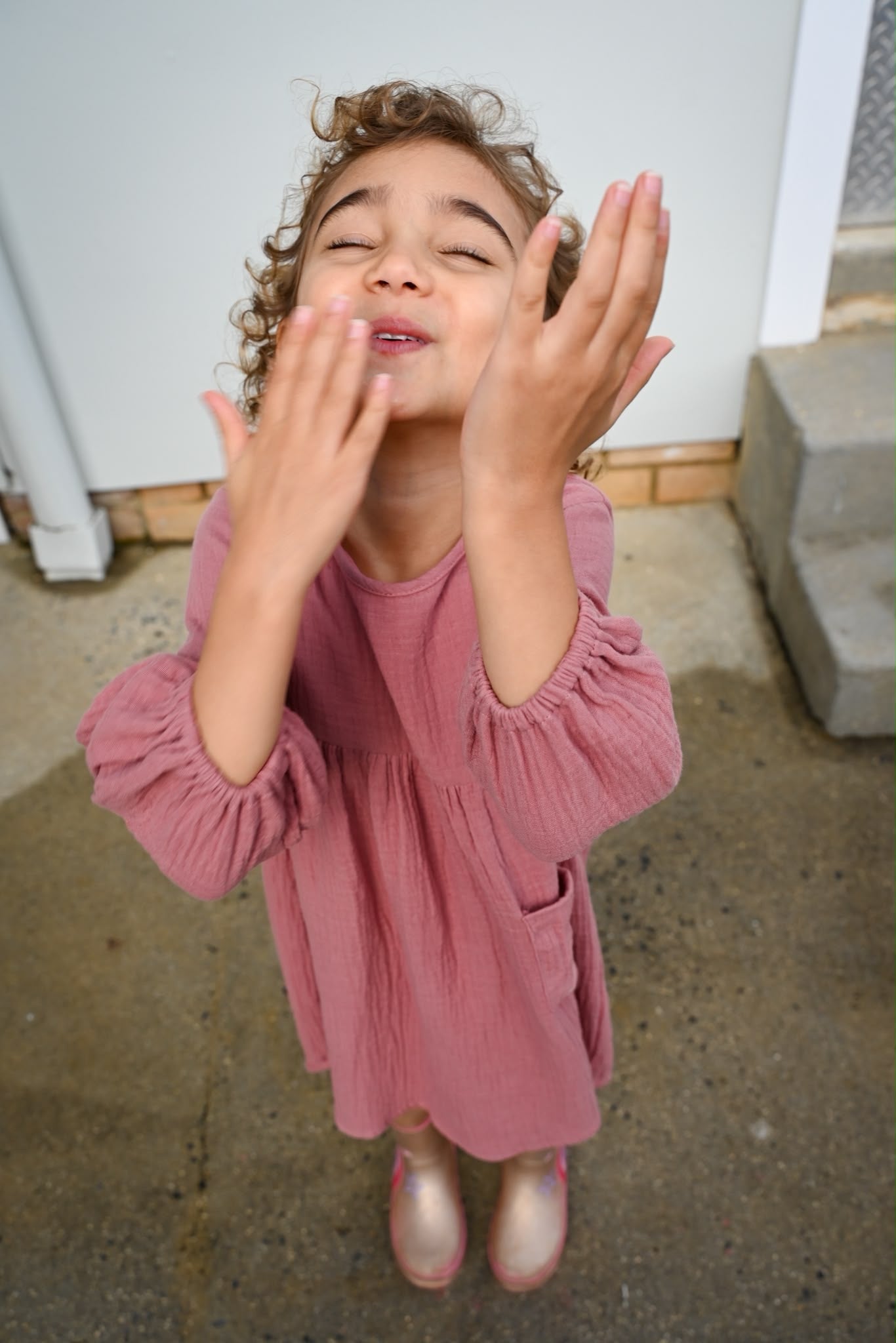 A young girl with curly brown hair, wearing a MilaMeli Evie Dress Dusty Rose Muslin Cotton Embroidered Feather Pocket.