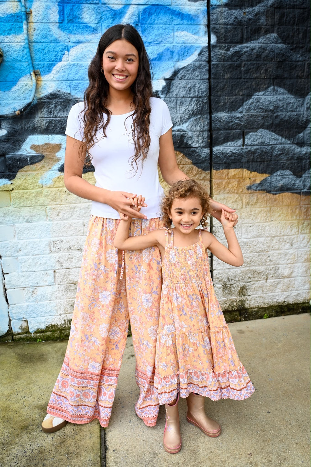 Lifestyle shot of a woman and child wearing matching Aria Spring Dress Sunrise Botanica Rayon, a bohemian-style clothing in soft peach-orange, dusty rose pink, and light lavender floral patterns, paired with a solid white crewneck top.