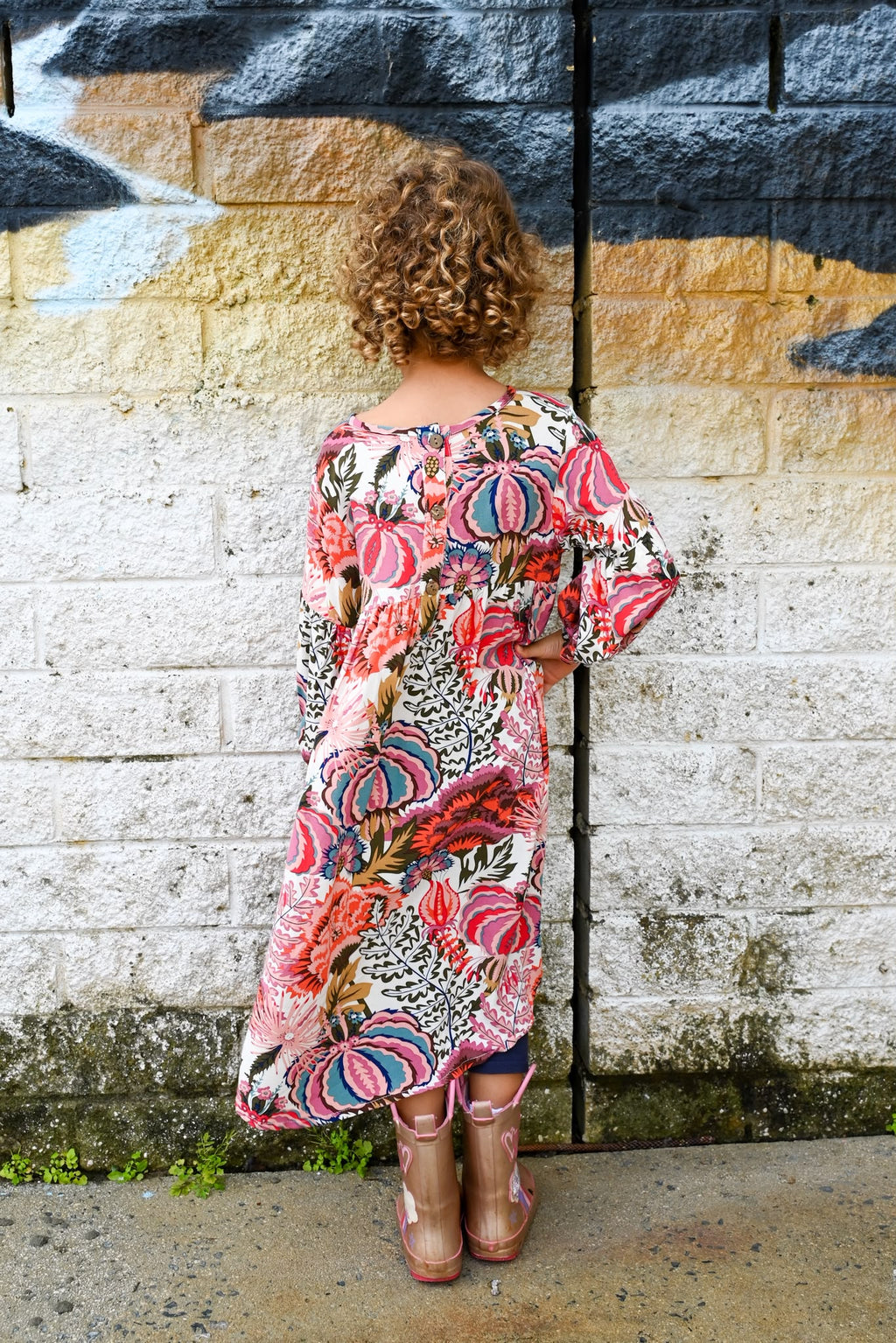 A woman with curly brown hair stands with her back to the camera, wearing a short-sleeve bohemian-style maxi dress Evie Dress Pink Agave Slub Rayon.