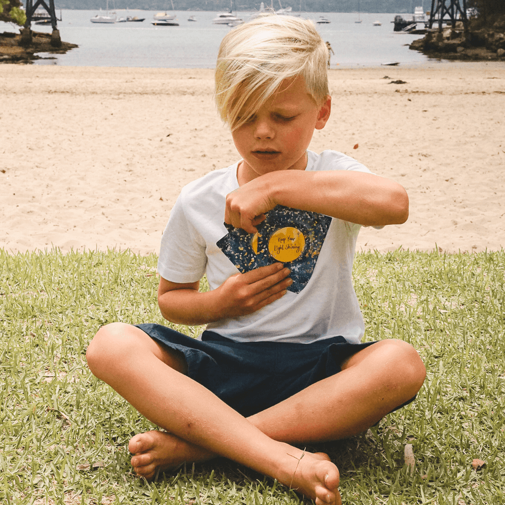 Young boy sitting on grass by a beach with boats in the background