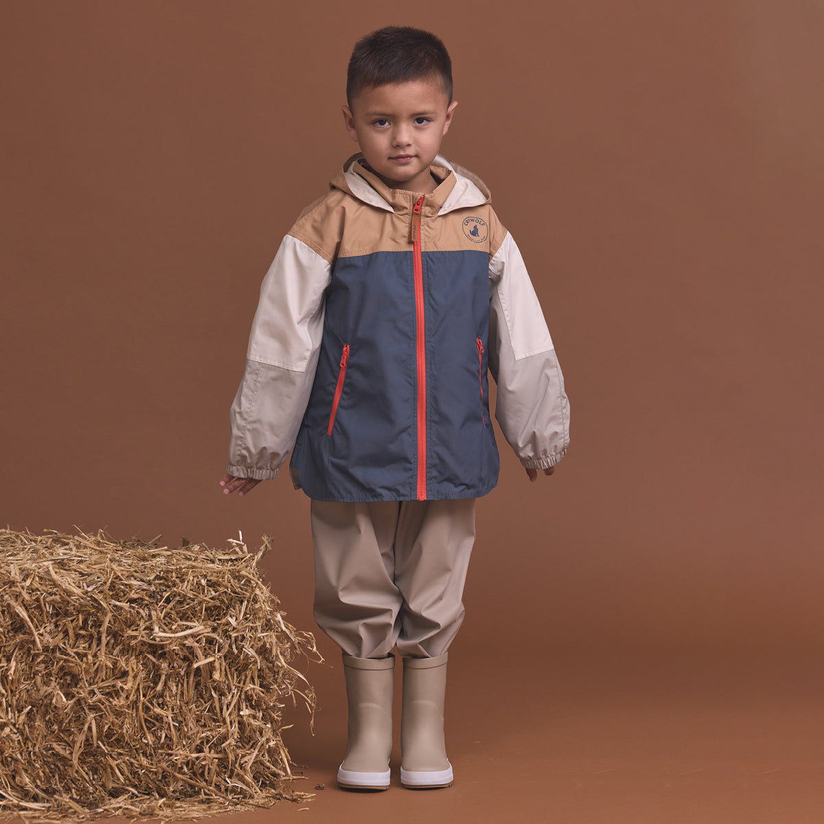 Child wearing the Crywolf Packable Jacket Ash Colourblock and pants standing next to a bale of hay on a brown background