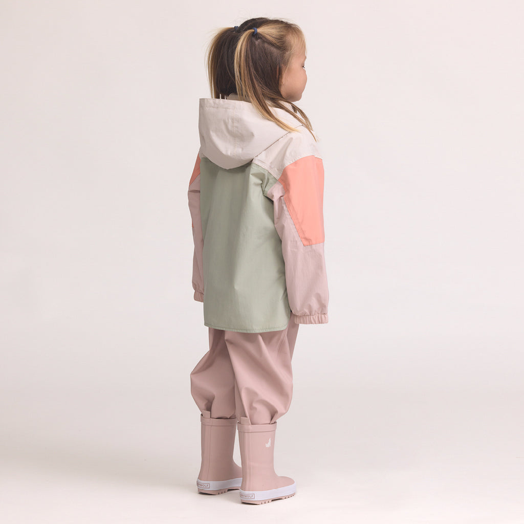 Child wearing a Color-blocked kids' waterproof jacket featuring an off-white upper body and hood, a sage green central torso, and terracotta pink sleeves with a cream zipper, pink pants and Crywolf Rain Boots Dusty Pink on a light background.