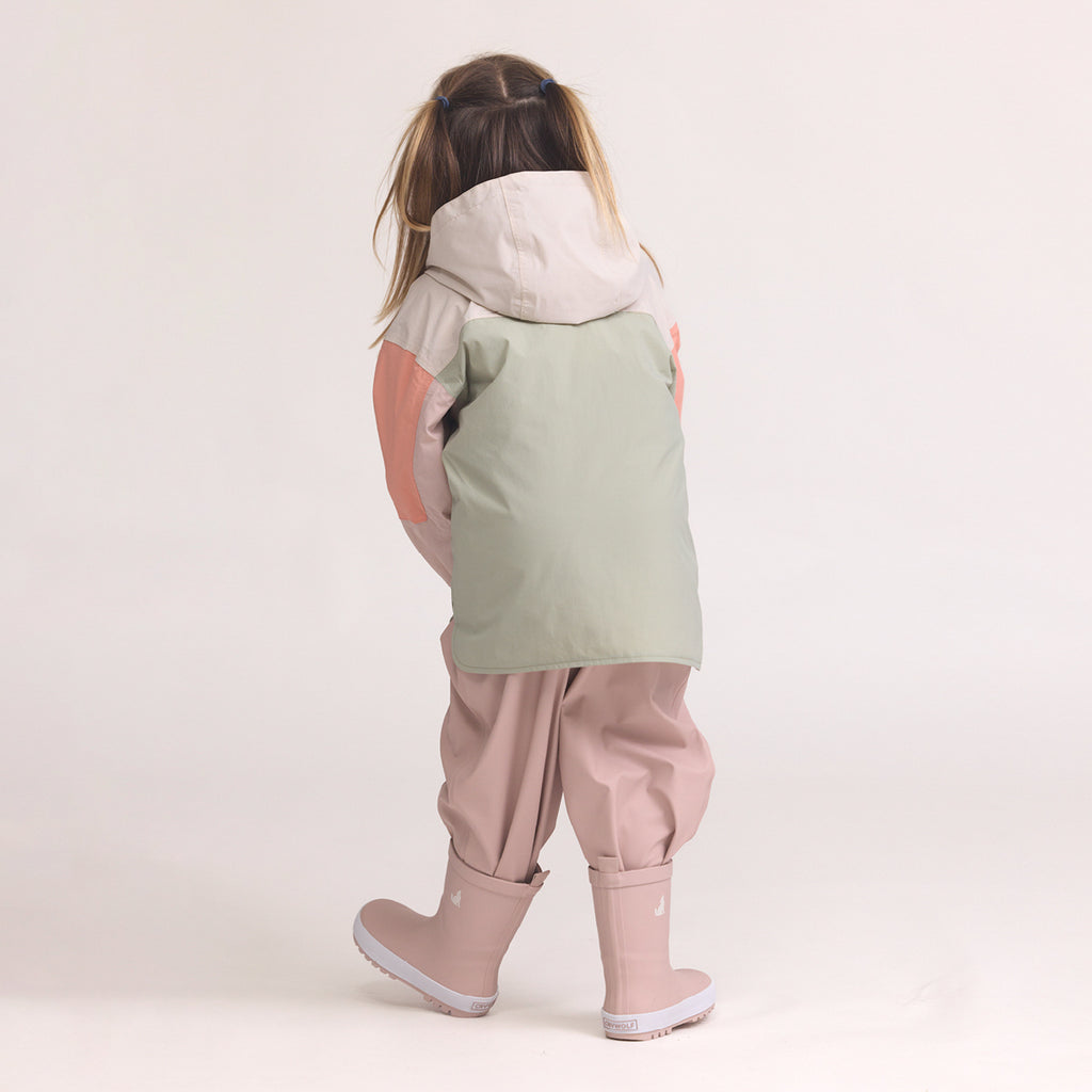 Child wearing a Color-blocked kids' waterproof jacket featuring an off-white upper body and hood, a sage green central torso, and terracotta pink sleeves with a cream zipper, pink pants and Crywolf Rain Boots Dusty Pink on a light background.