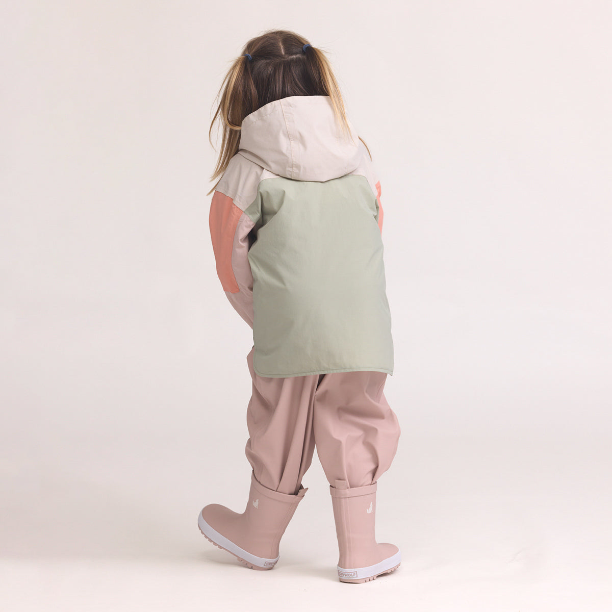 Child wearing a Color-blocked kids' waterproof jacket featuring an off-white upper body and hood, a sage green central torso, and terracotta pink sleeves with a cream zipper, pink pants and Crywolf Rain Boots Dusty Pink on a light background.
