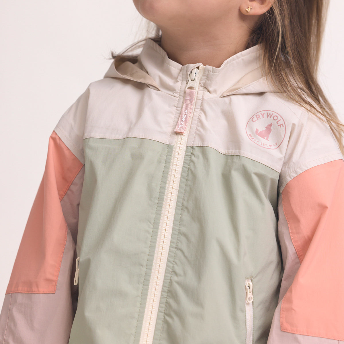 Color-blocked kids' waterproof jacket featuring an off-white upper body and hood, a sage green central torso, and terracotta pink sleeves with a cream zipper.