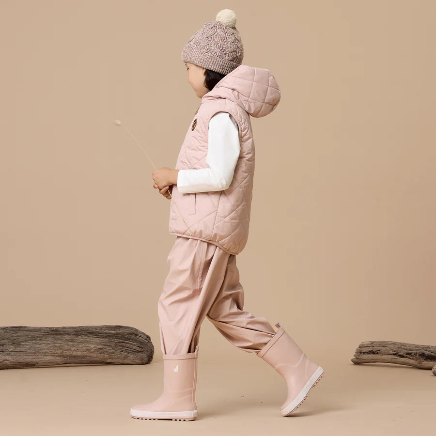 Child wearing a pink winter outfit with a vest, pants, and Crywolf Rain Boots on a beige background