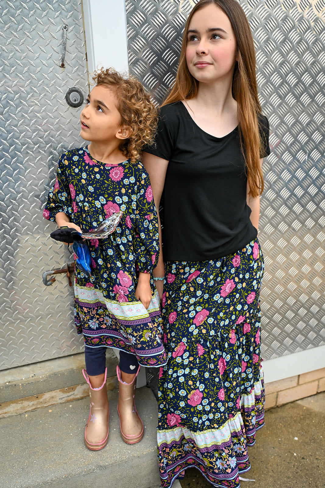 Two girls standing outdoors. The girl on the right wears a black t-shirt and a long maxi Indigo Wildflower skirt, a colorful floral print, while the girl in the left wears Lotte Dress Indigo Wildflower top.