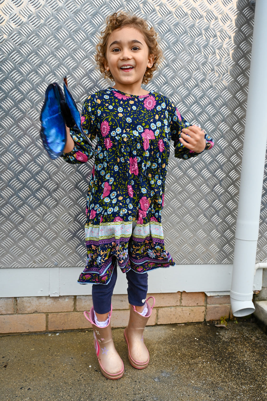 A young girl with curly hair stands outdoors, wearing a Lotte Dress Indigo Wildflower top.