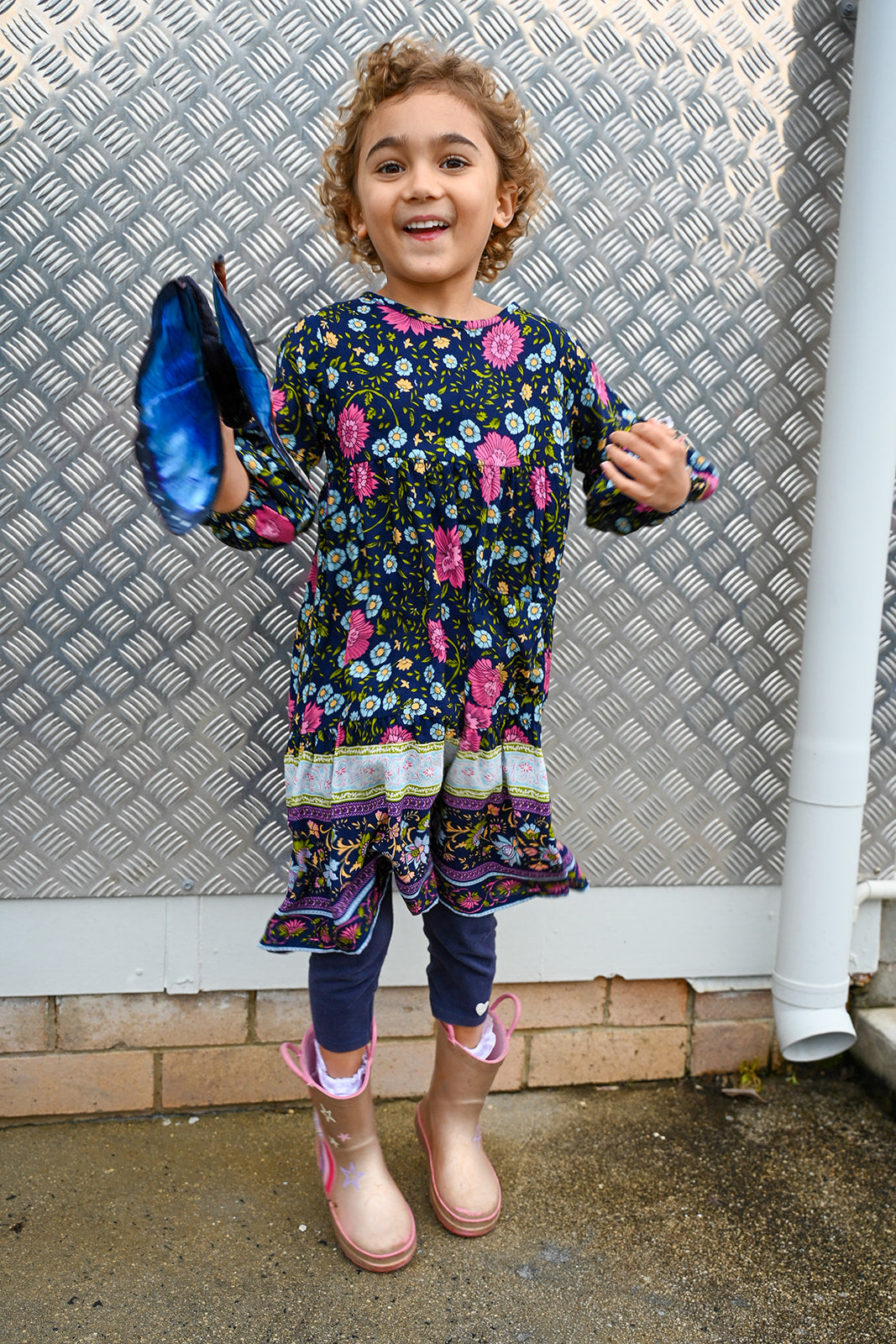 A young girl with curly hair stands outdoors, wearing a Lotte Dress Indigo Wildflower top.