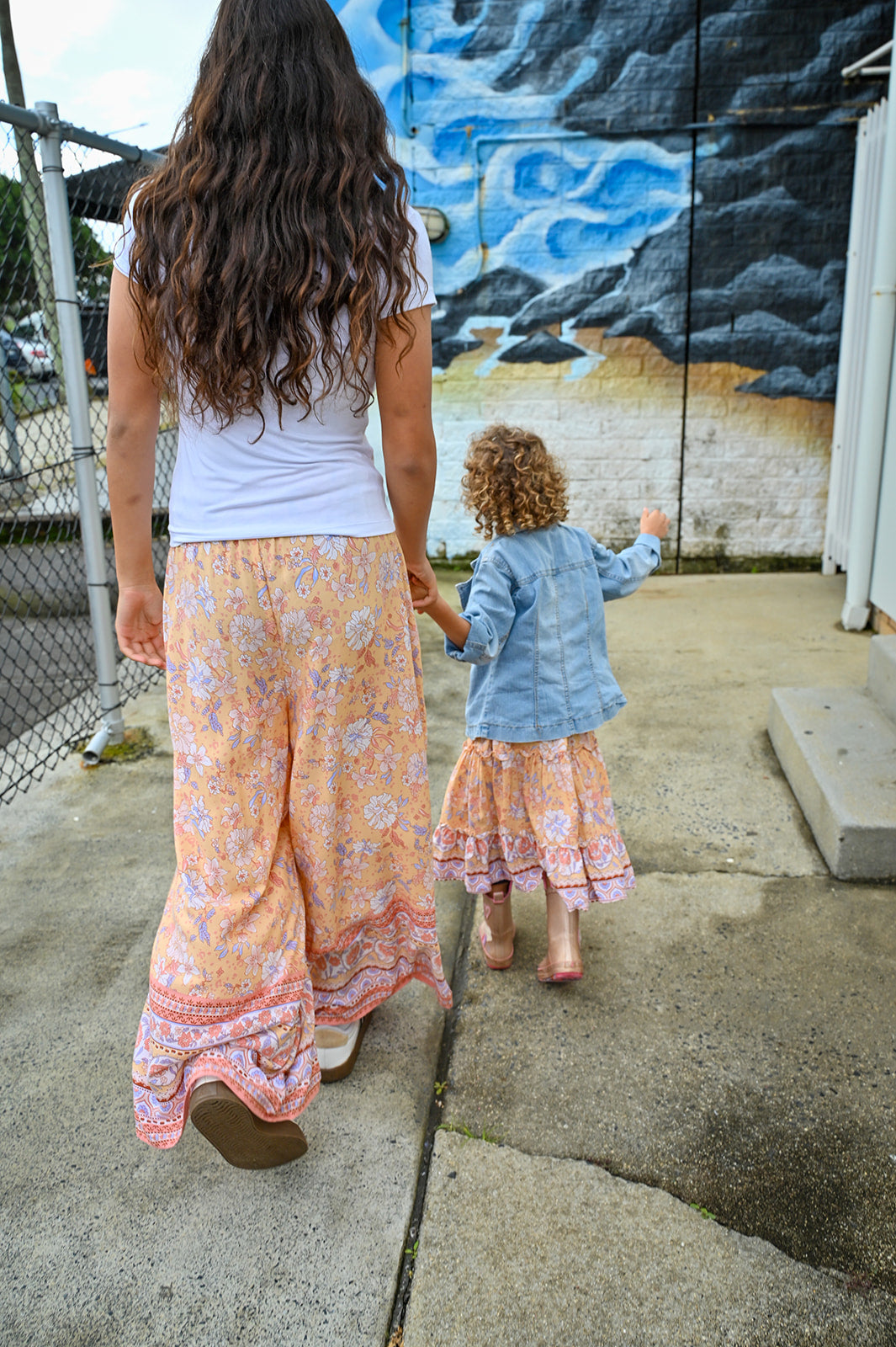 A woman and a young child walking hand in hand outdoors. The woman wears a white top and Mila Meli Lounge Pants Sunrise Botanica.