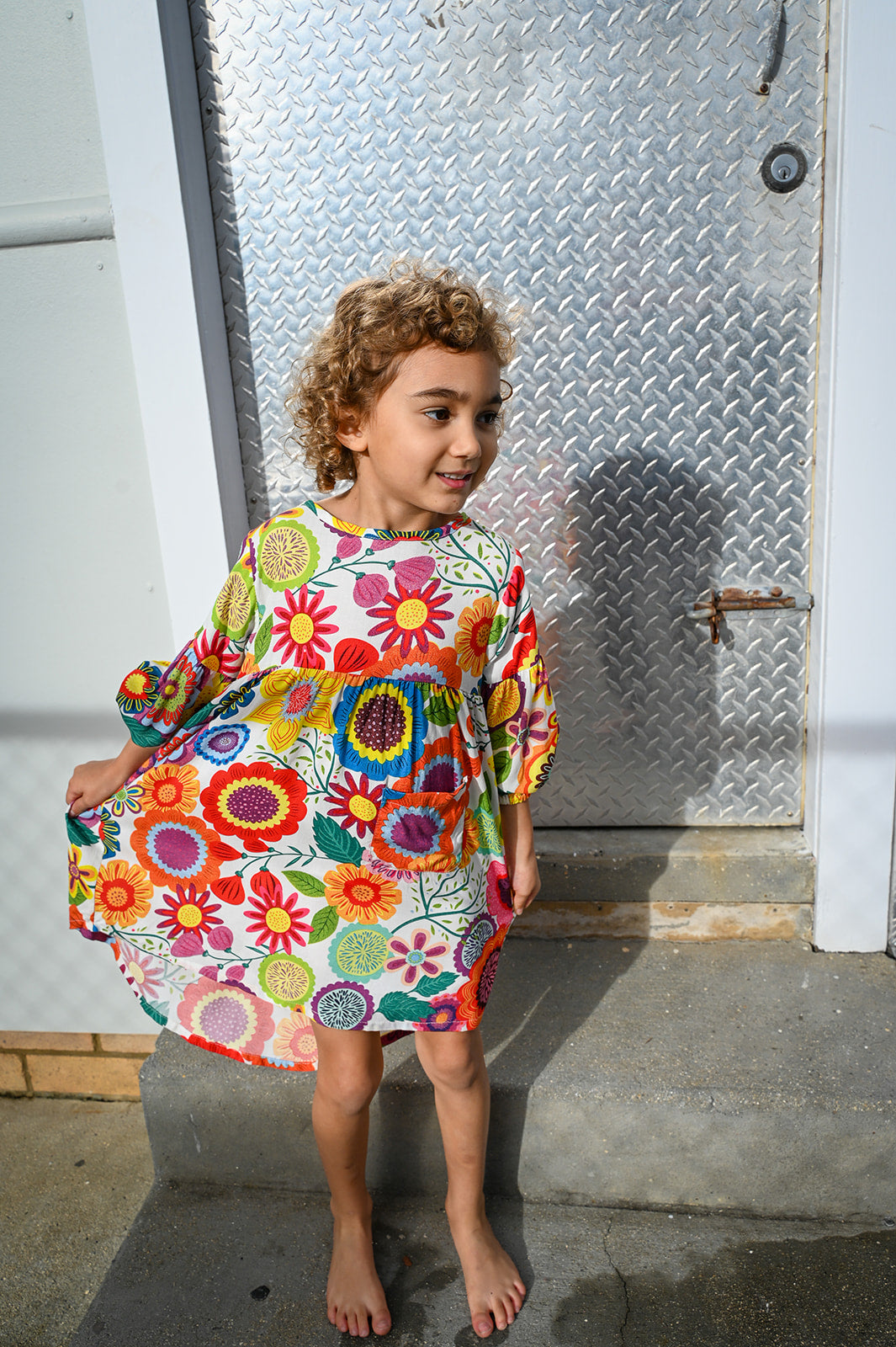 A young girl with curly brown hair stands barefoot against a weathered concrete wall, wearing a Evie Dress Flower Pop Brights.