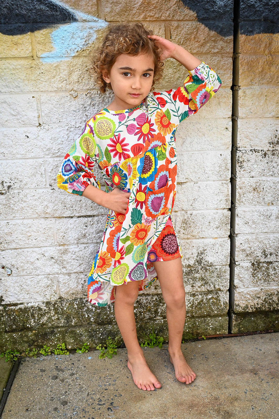 A young girl with curly brown hair stands barefoot against a weathered concrete wall, wearing a Evie Dress Flower Pop Brights.