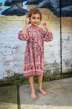 A young girl with curly brown hair stands barefoot, wearing a pink and red patterned long-sleeve tunic dress, Lottie Dress Blush Wildflower Print.