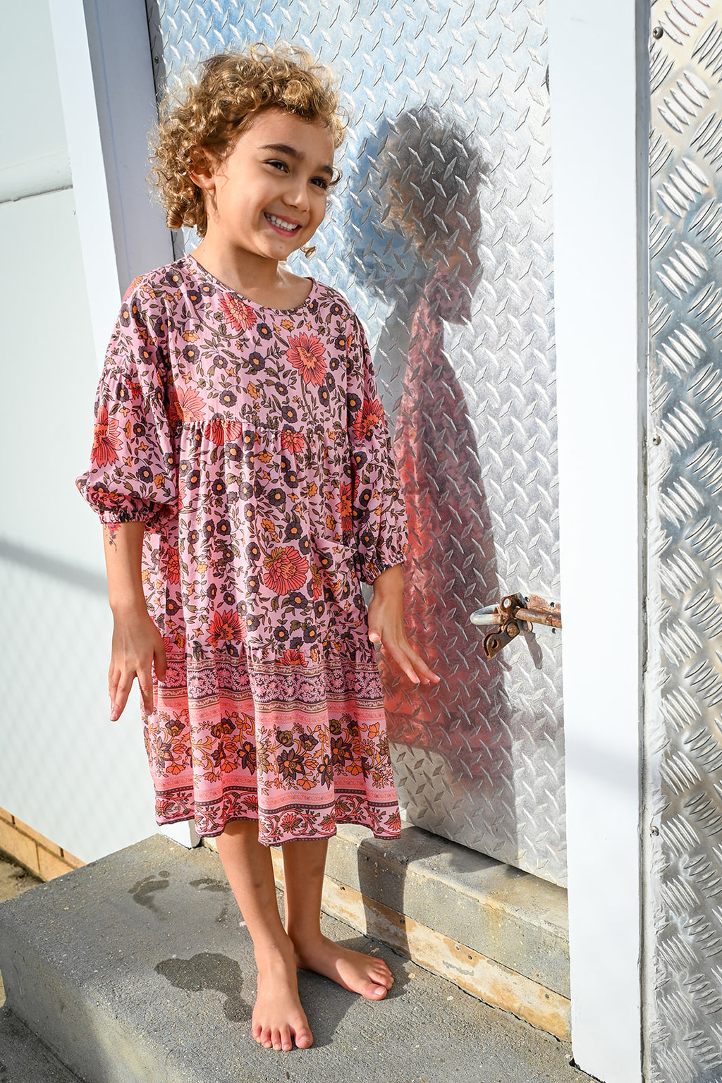 A young girl with curly brown hair stands barefoot, wearing a pink and red patterned long-sleeve tunic dress, Lottie Dress Blush Wildflower Print.