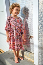 A young girl with curly brown hair stands barefoot, wearing a pink and red patterned long-sleeve tunic dress, Lottie Dress Blush Wildflower Print.