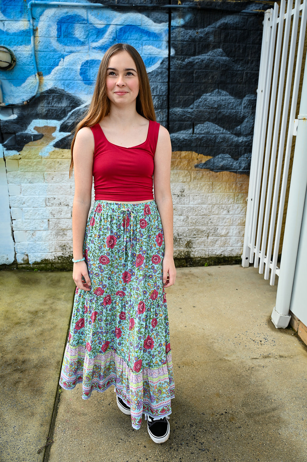 A young woman stands outdoors in front of a painted brick wall, wearing a sleeveless red tank top and a long, Maeve Maxi Skirt Aqua Wildflower.