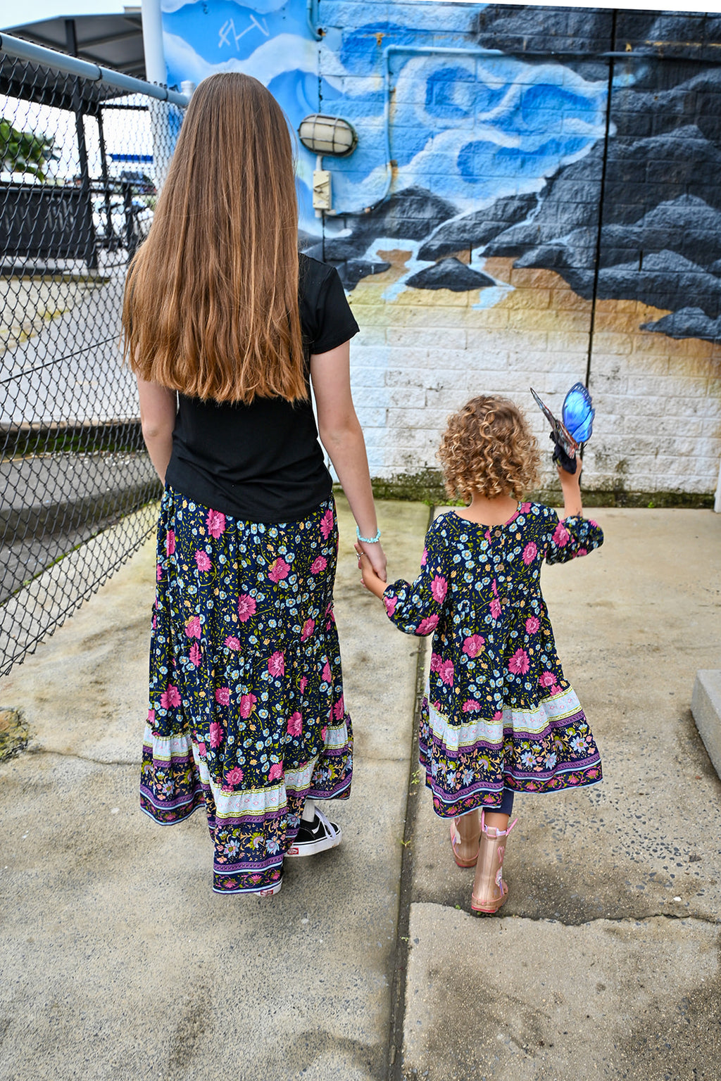 Two girls standing outdoors. The girl on the left wears a black t-shirt and a long maxi Indigo Wildflower skirt, a colorful floral print, while the girl in the right wears Lotte Dress Indigo Wildflower top.
