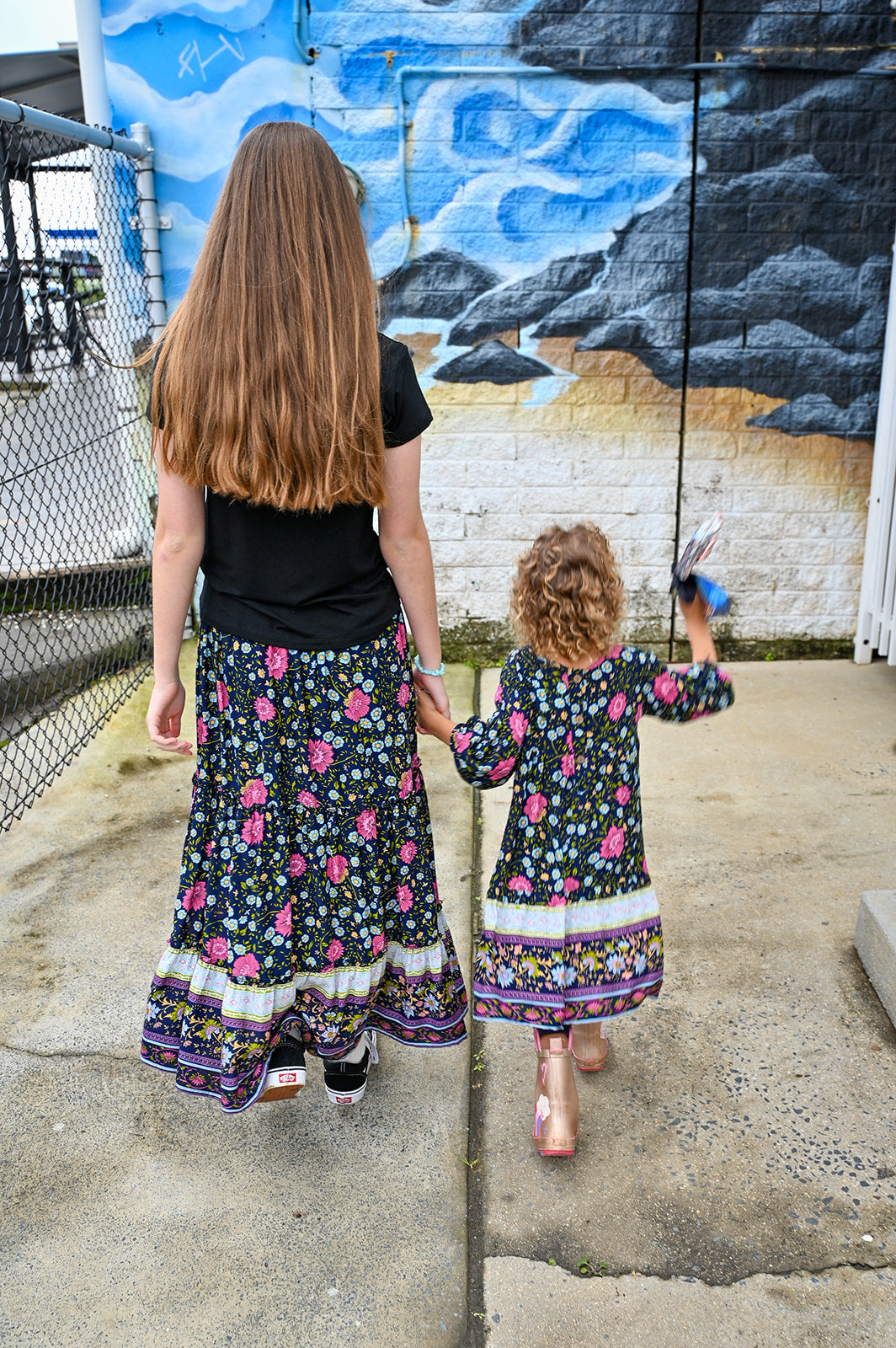 Two girls standing outdoors. The girl on the left wears a black t-shirt and a long maxi Indigo Wildflower skirt, a colorful floral print, while the girl in the right wears Lotte Dress Indigo Wildflower top.