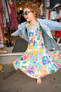  A child twirling in Aria Spring Dress Sweet Soul Rayon Print, a vibrant multi-colored patterned tiered dress with bright graphics, worn with an open denim jacket and colorful sunglasses.