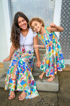 A woman and a young girl sitting together on concrete steps, wearing matching Aria Spring Dress Sweet Soul Rayon Print, a vibrant multi-colored patterned tiered dress with bright graphics.