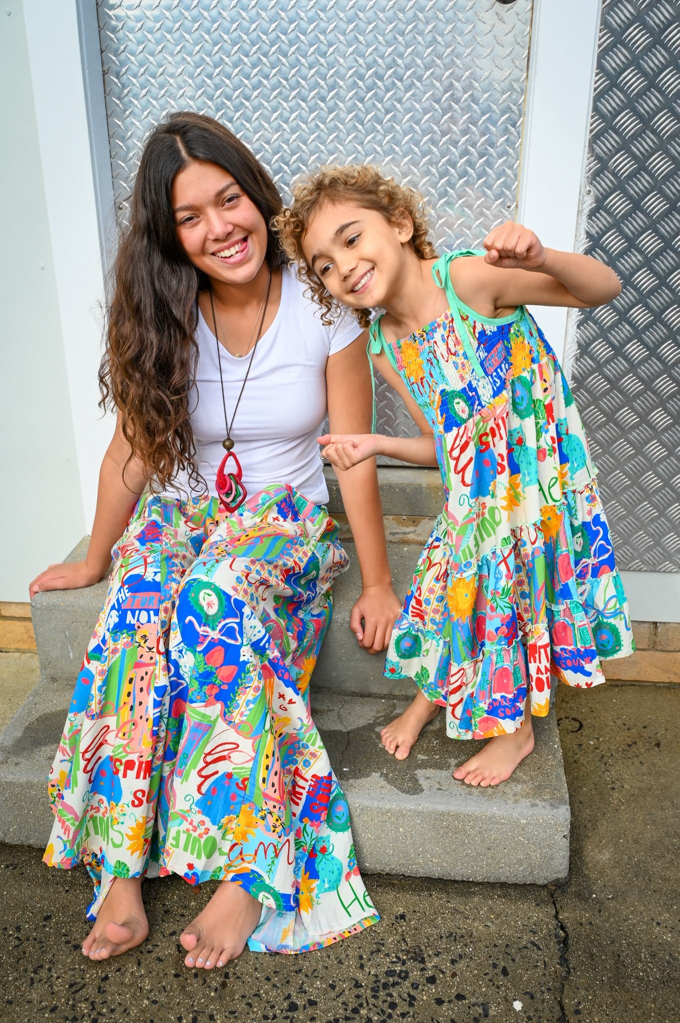 A woman and a young girl sitting together on concrete steps, wearing matching colorful abstract-patterned maxi dresses and long skirts in Byron  Sweet SoulPrint.