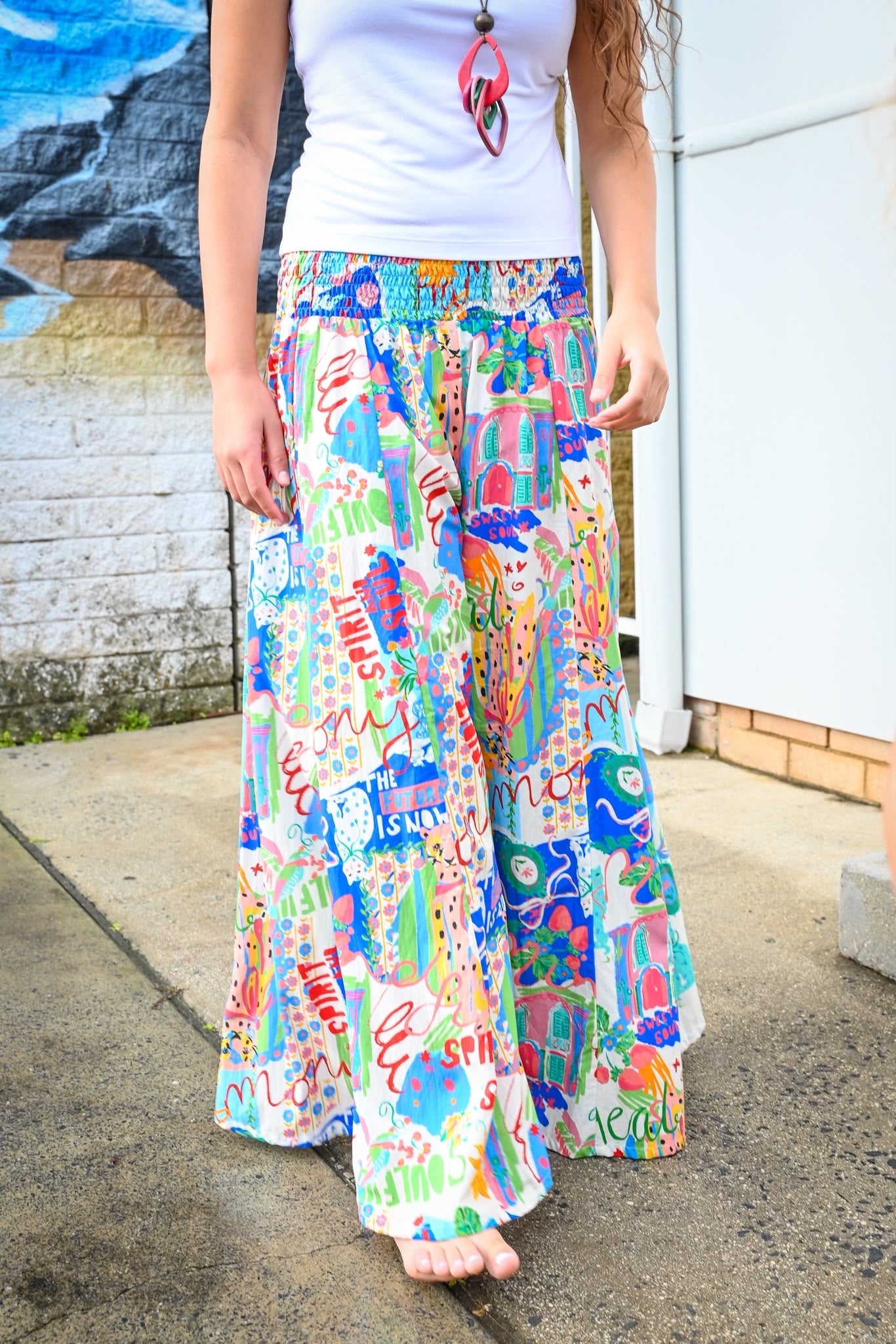 A person wearing a white tank top and very wide-leg, multicolored patchwork-print maxi pants Byron Pant Sweet SoulPrint.