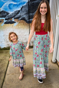 Two girls walking hand in hand outdoors. The older girl wears a red tank top and a Maeve Maxi Skirt Aqua Wildflower Print, while the younger girl wears a matching Aqua Wildflower Print Lottie dress.
