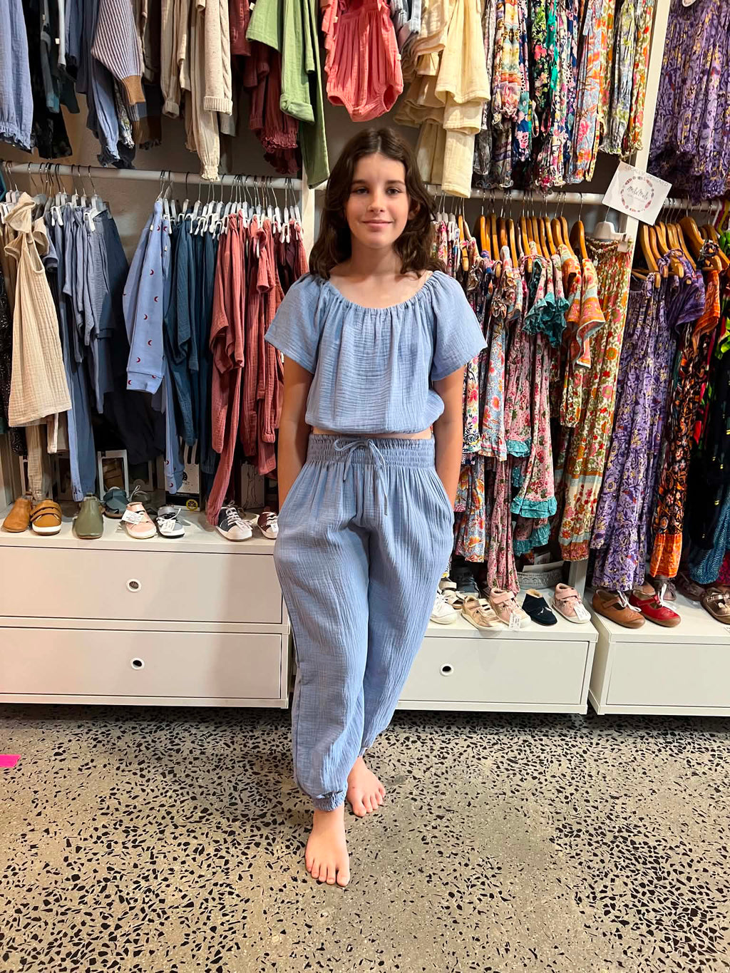 Young girl in a blue outfit standing in a clothing store wearing MilaMeli Lil Watego Pants, a Light blue textured double-gauze short-sleeve jumpsuit featuring a smocked elastic waistband with a decorative drawstring and cinched ankles.