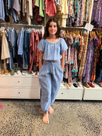 Young girl in a blue outfit standing in a clothing store wearing MilaMeli Lil Watego Pants, a Light blue textured double-gauze short-sleeve jumpsuit featuring a smocked elastic waistband with a decorative drawstring and cinched ankles.