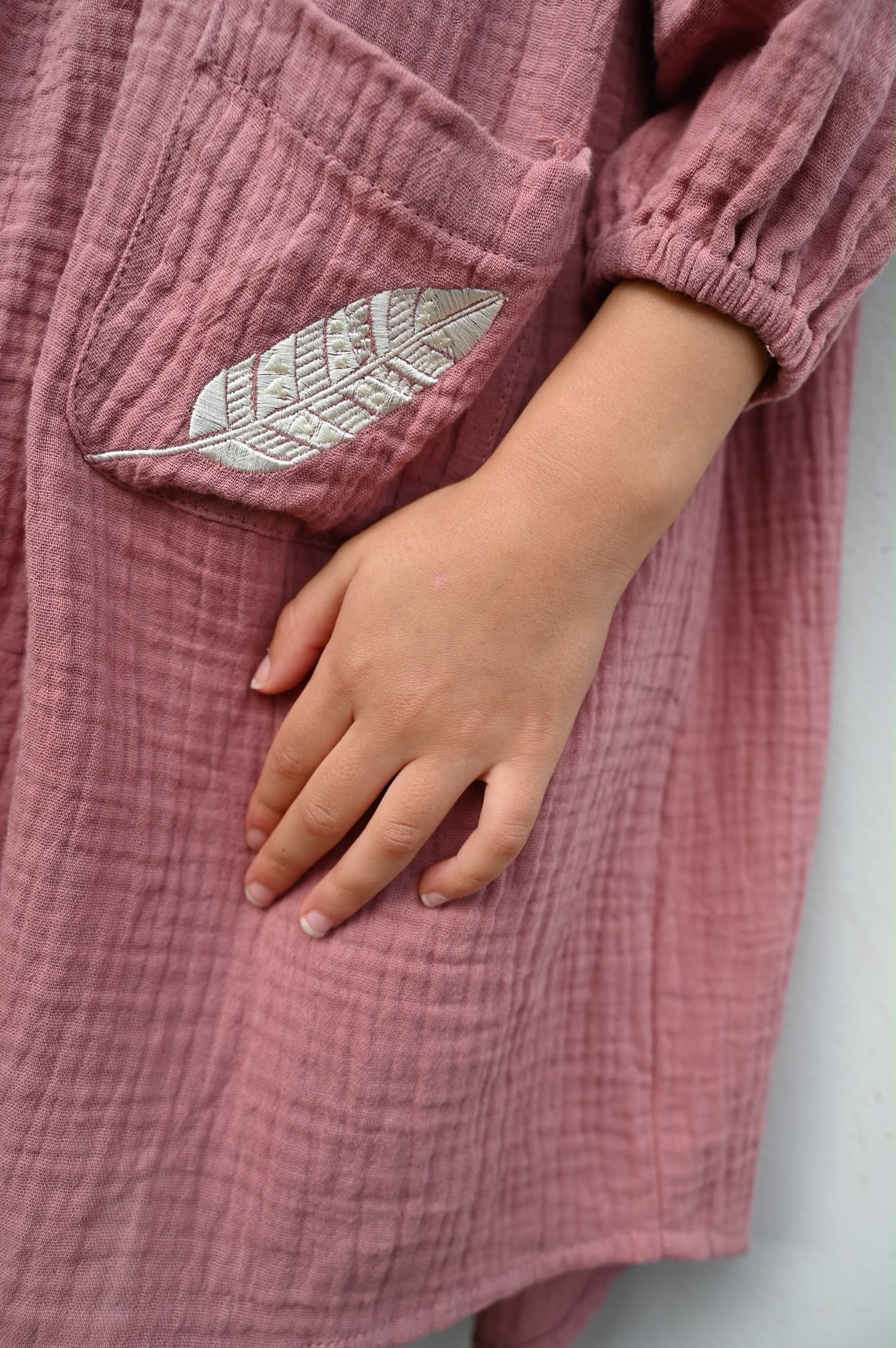 Close-up of MilaMeli Evie Dress Dusty Rose Muslin Cotton Embroidered Feather Pocket, worn by a child with one hand resting on the fabric.